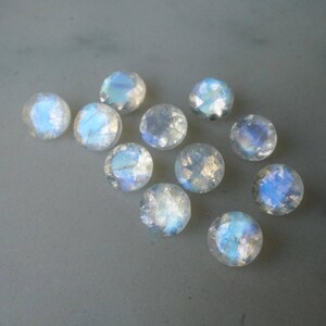 May include: A collection of iridescent, round gemstones with a faceted cut. The stones display a range of colors, including blues and whites, and are scattered on a light gray surface. These gemstones are likely moonstones.