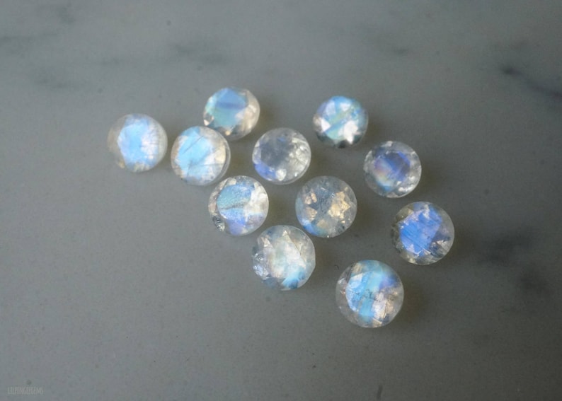 May include: A collection of iridescent, round gemstones with a faceted cut. The stones display a range of colors, including blues and whites, and are scattered on a light gray surface. These gemstones are likely moonstones.