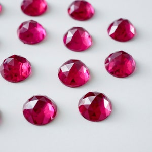 Puede incluir: Una colección de 12 gemas facetadas de color rosa brillante dispuestas en filas sobre una superficie blanca.