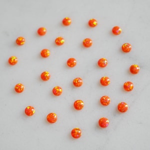 May include: A collection of 25 small, round, orange opal cabochons. The cabochons are scattered on a white surface.