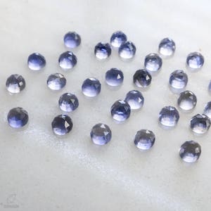 May include: A collection of 25 faceted, iridescent, purple-blue gemstones. The stones are scattered on a white surface.