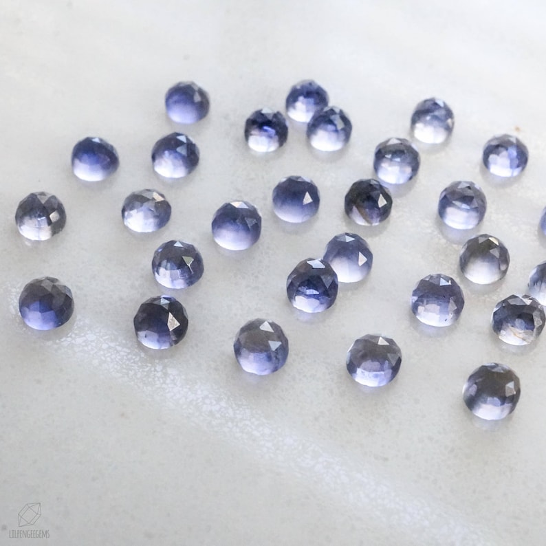 May include: A collection of 25 faceted, iridescent, purple-blue gemstones. The stones are scattered on a white surface.