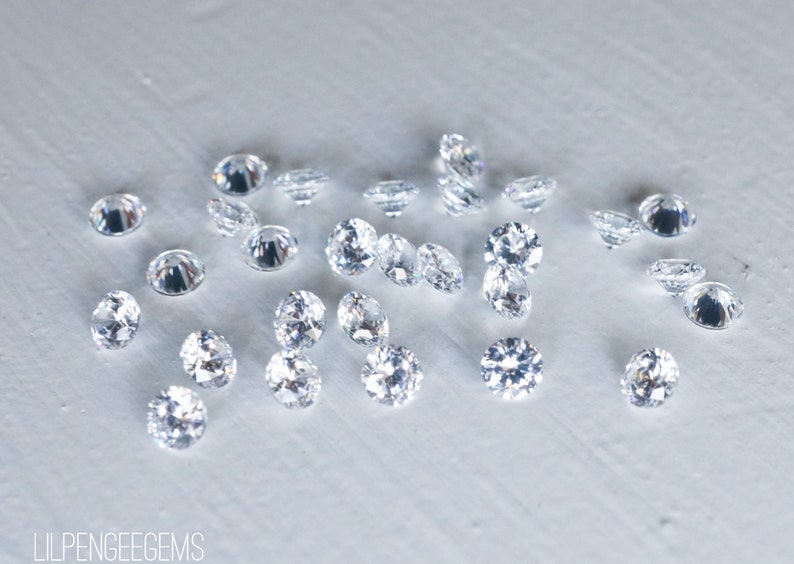 May include: A collection of clear, round, faceted gemstones, scattered on a white surface.