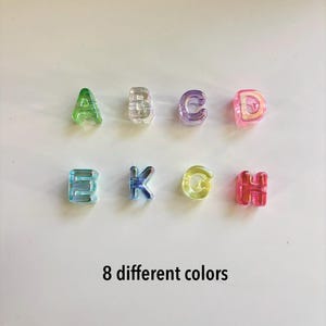 Puede incluir: Ocho cuentas de alfabeto de plástico translúcido dispuestas en dos filas, con las letras de la A a la H en colores distintos que incluyen verde, transparente, morado, rosa, turquesa, azul, amarillo y rojo, con el texto 8 different colors.