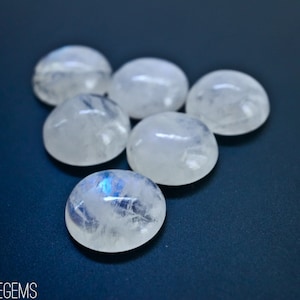 Puede incluir: Seis cabujones lisos de piedra lunar blanca con destellos iridiscentes azules y morados. Las piedras están dispuestas en forma triangular sobre un fondo azul oscuro.
