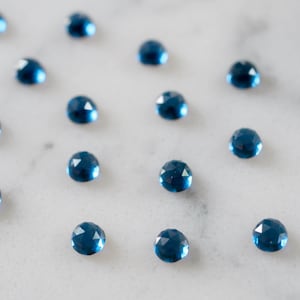 May include: A collection of 16 blue faceted gemstones arranged in rows on a white marble surface.