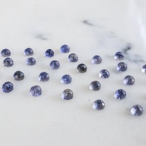 May include: A collection of 20 small, round, faceted gemstones. The gemstones are a light blue color with a hint of purple. They are arranged in a scattered pattern on a white surface.