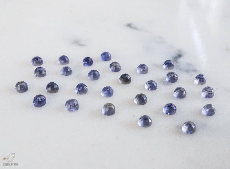 May include: A collection of 20 small, round, faceted gemstones. The gemstones are a light blue color with a hint of purple. They are arranged in a scattered pattern on a white surface.