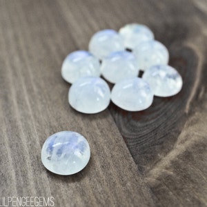 Puede incluir: Un grupo de 9 cabochones de piedra lunar lisos y blancos, con un solo caboch&oacute;n en primer plano. Los cabochones est&aacute;n dispuestos en un grupo suelto sobre una superficie de madera.