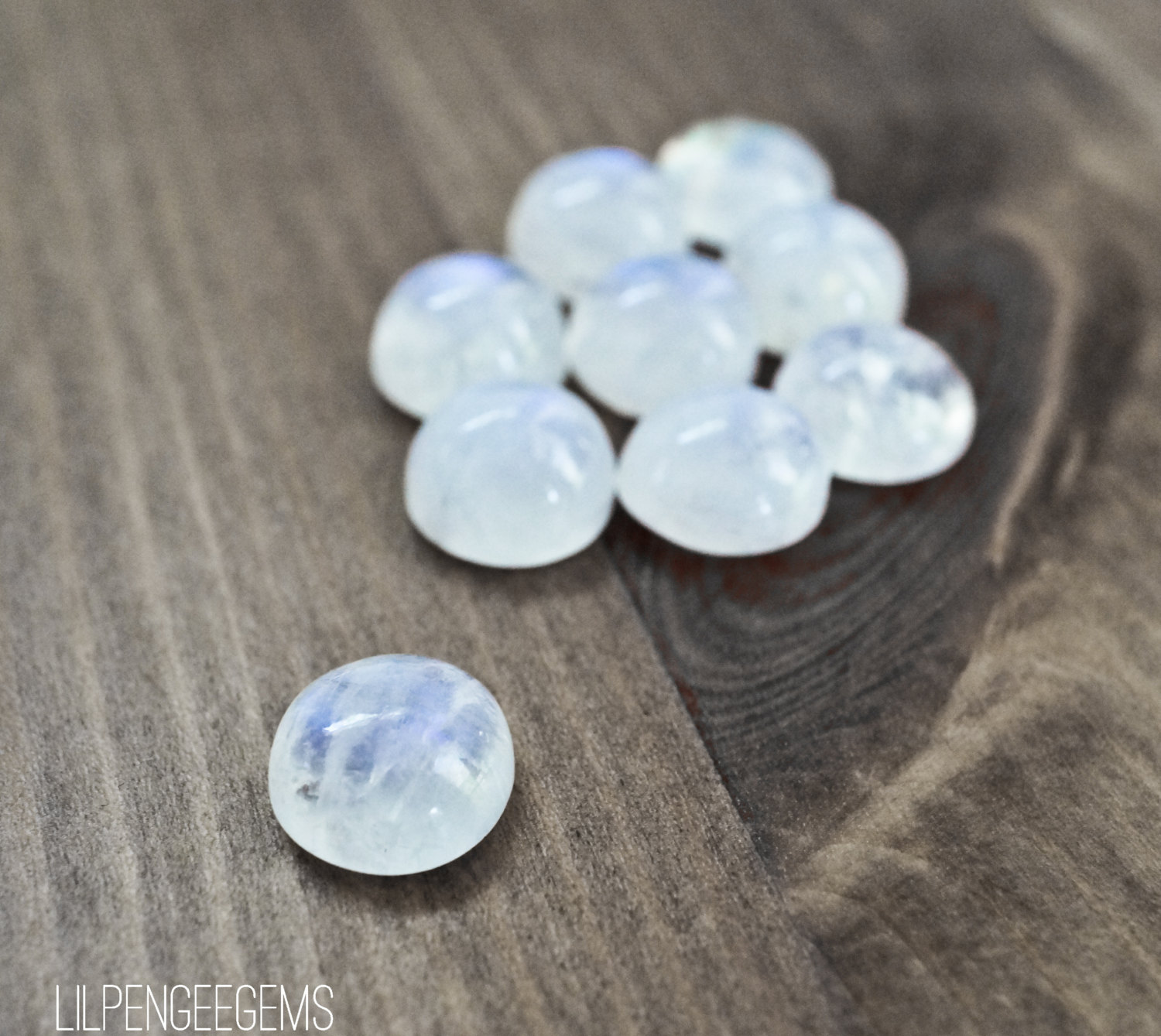 Puede incluir: Un grupo de 9 cabochones de piedra lunar lisos y blancos, con un solo caboch&oacute;n en primer plano. Los cabochones est&aacute;n dispuestos en un grupo suelto sobre una superficie de madera.