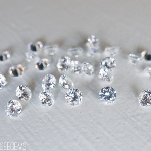 May include: A close-up of a group of clear, round, faceted gemstones on a white surface. The gemstones are scattered randomly and appear to be loose.