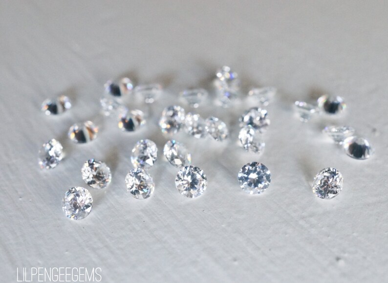 May include: A close-up of a group of clear, round, faceted gemstones on a white surface. The gemstones are scattered randomly and appear to be loose.