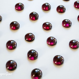 May include: A collection of 20 round, cabochon-cut, deep red gemstones arranged in a pattern on a white surface.