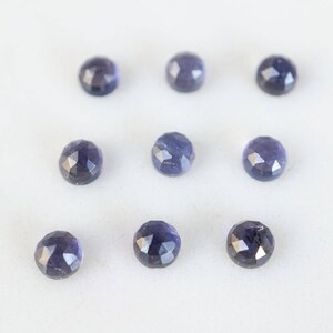May include: Ten faceted, round, dark blue gemstones on a white surface.
