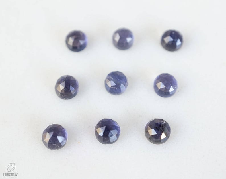 May include: Ten faceted, round, dark blue gemstones on a white surface.