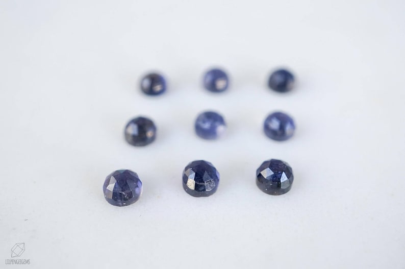 May include: Nine round, faceted, dark blue gemstones on a white surface.