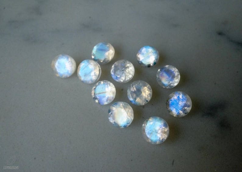 May include: A collection of round, iridescent moonstone cabochons. Each stone displays a milky white base with flashes of blue and rainbow hues. The gemstones are scattered on a gray surface, showcasing their polished, domed shape.