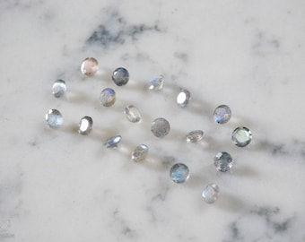 May include: A collection of round, iridescent labradorite gemstones in various shades of blue and grey, scattered on a white marble surface.