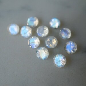 May include: A collection of round, iridescent moonstone cabochons. The gemstones display a range of colors, including blue, white, and hints of yellow. The stones are scattered on a light gray surface.