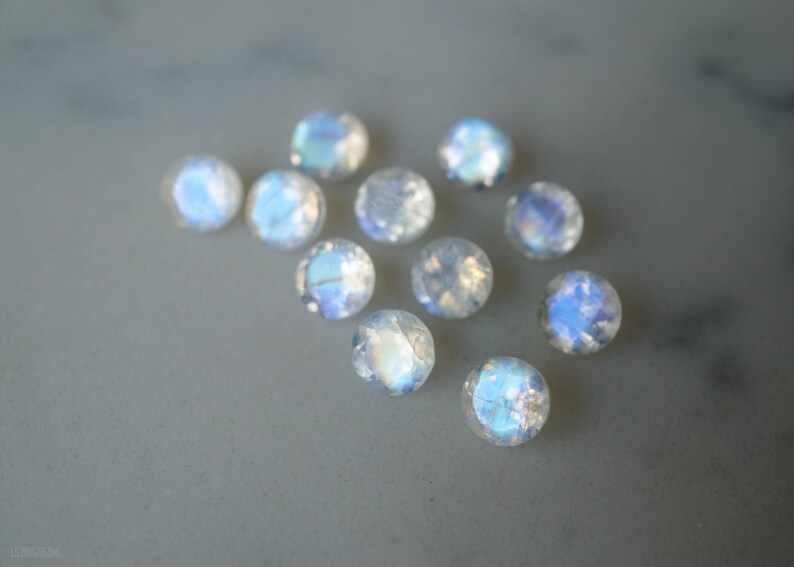 May include: A collection of round, iridescent moonstone cabochons. The gemstones display a range of colors, including blue, white, and hints of yellow. The stones are scattered on a light gray surface.