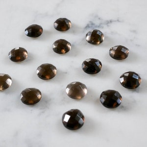 Puede incluir: Una colección de 15 cabujones de cuarzo ahumado facetados, cada uno con un color marrón oscuro y una superficie lisa y pulida. Las piedras están dispuestas en una cuadrícula sobre un fondo de mármol blanco.