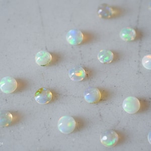 May include: A collection of 14 iridescent white opal cabochons, scattered on a light gray surface. The opals are round and have a smooth, polished finish.