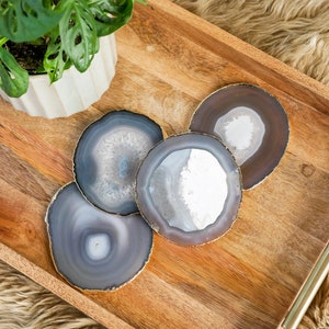 May include: Four agate coasters with gold edges are arranged on a wooden tray. The coasters have a natural, layered appearance with shades of gray, white, and brown. A potted plant with green leaves is in the background.