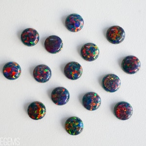 May include: A collection of 15 round, black opal cabochons with iridescent colors, including red, green, blue, and yellow. The opals are arranged in a scattered pattern on a white background.