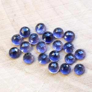 May include: A collection of 20 faceted amethyst gemstones, each with a deep purple hue and a slightly translucent appearance. The stones are arranged in a loose cluster on a light wood surface.