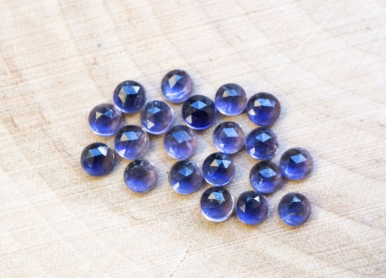 May include: A collection of 20 faceted amethyst gemstones, each with a deep purple hue and a slightly translucent appearance. The stones are arranged in a loose cluster on a light wood surface.