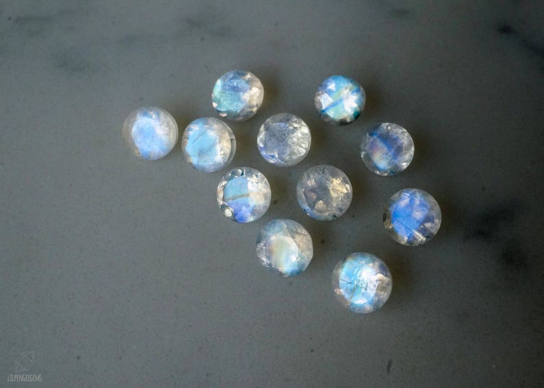May include: A collection of round, faceted moonstone gemstones. The stones display a shimmering, iridescent play of color, with hues of blue, white, and hints of other colors. The gemstones are scattered on a gray surface.
