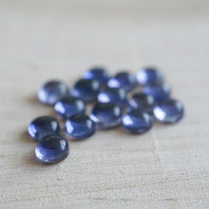 May include: A group of 15 small, round, blue gemstones. The gemstones are smooth and shiny, and they are arranged in a loose cluster on a light wood surface.