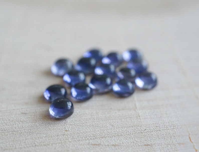 May include: A group of 15 small, round, blue gemstones. The gemstones are smooth and shiny, and they are arranged in a loose cluster on a light wood surface.