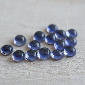 May include: A group of 15 round, smooth, blue-grey gemstones. The gemstones are arranged in a loose cluster on a light wood surface.