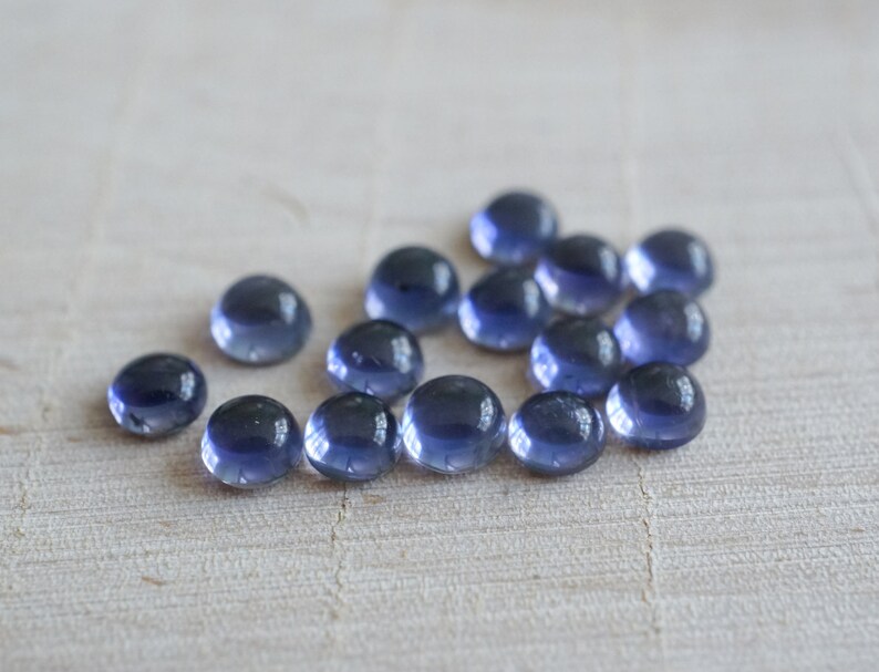 May include: A group of 15 round, smooth, blue-grey gemstones. The gemstones are arranged in a loose cluster on a light wood surface.