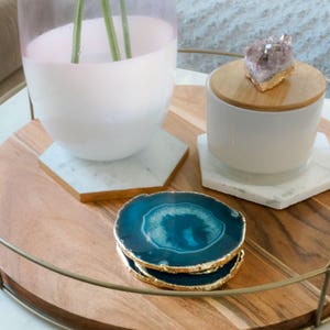 May include: Two teal agate coasters with gold trim on a wooden tray with a white vase and a white ceramic container with a wooden lid and a purple crystal on top.