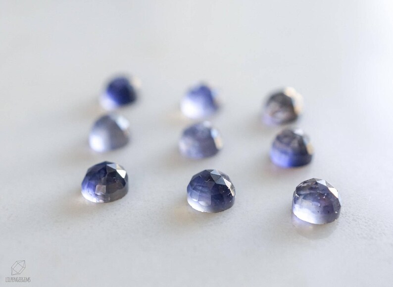 May include: A collection of eight faceted, blue-grey gemstones on a white surface. The gemstones are all approximately the same size and shape.