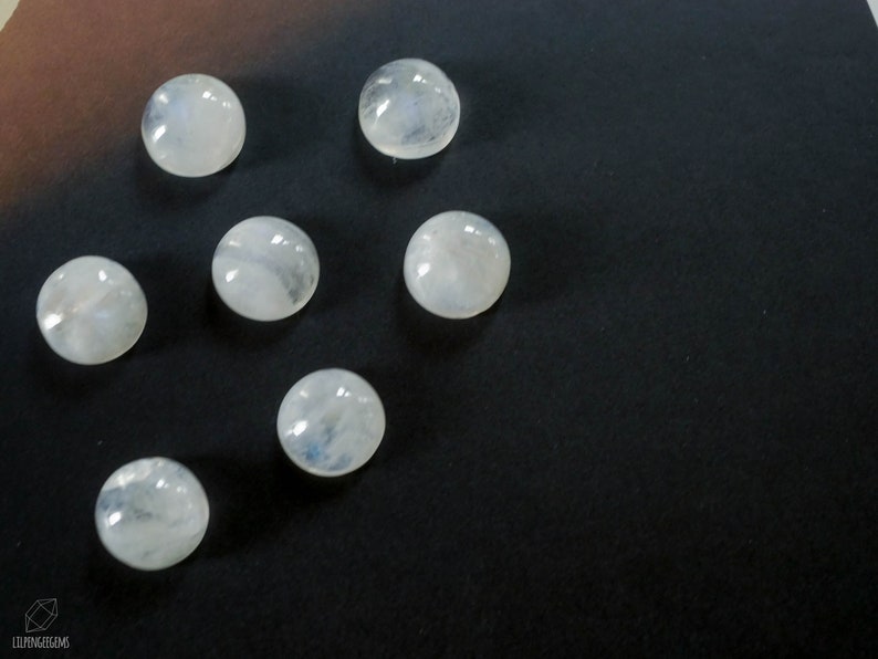 Puede incluir: Siete cabujones de piedra lunar blanca sobre un fondo negro. Las piedras son redondas y tienen una superficie lisa y pulida.