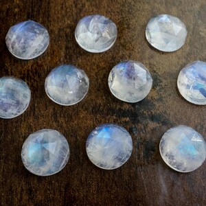 Puede incluir: Diez cabujones de piedra lunar iridiscente, cada uno con un patrón único de azul y blanco, están dispuestos sobre una superficie de madera marrón oscuro.