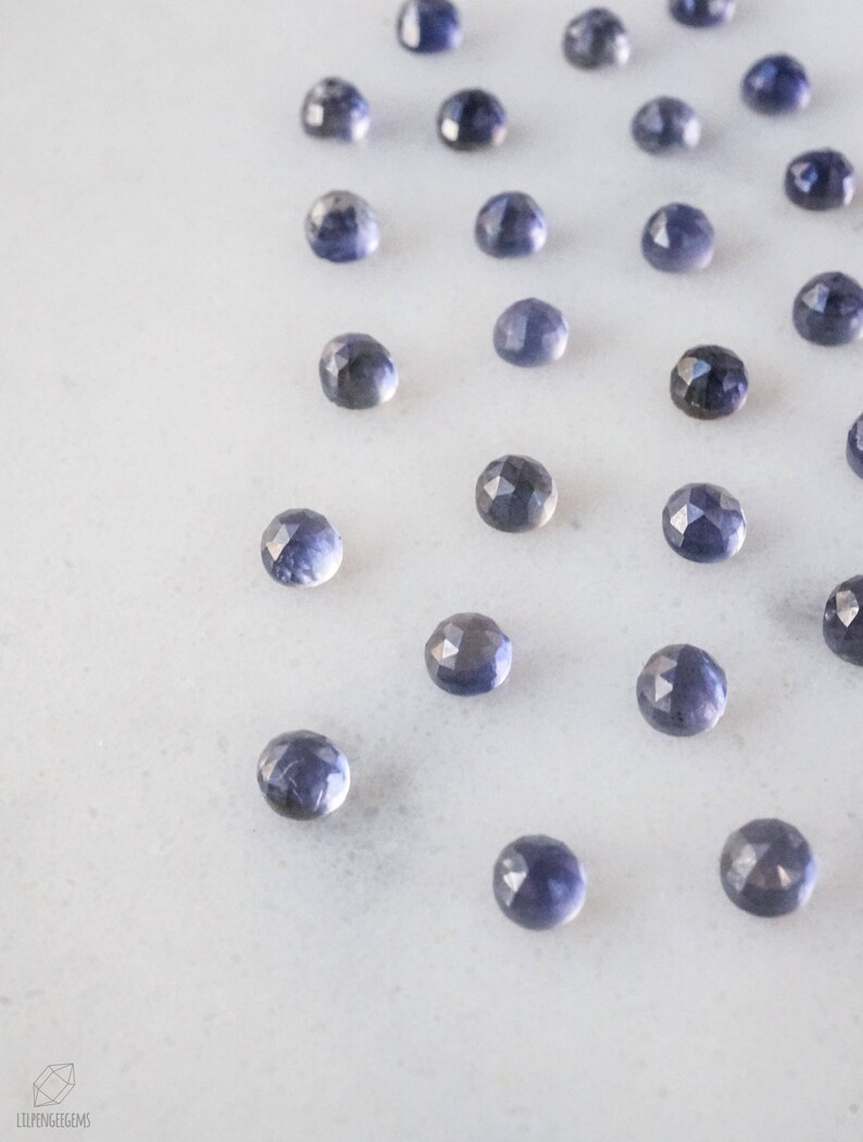 May include: A collection of 20 faceted, round, blue gemstones on a white surface.