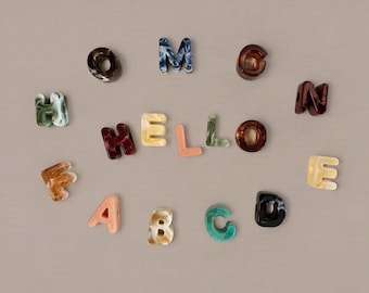 May include: A collection of individual alphabet letters made from marbled acrylic resin in various colors including brown, blue, green, orange, and cream. The letters are arranged in a circular pattern with the word HELLO spelled out in the center.