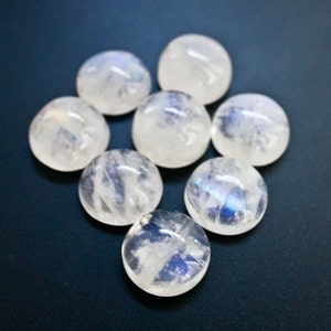 Puede incluir: Siete cabujones redondos de piedra lunar blanca con destellos iridiscentes azules y blancos. Las piedras est&aacute;n dispuestas en un c&iacute;rculo sobre un fondo azul oscuro.