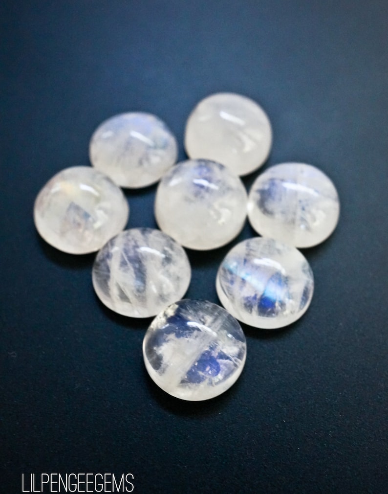 Puede incluir: Siete cabujones redondos de piedra lunar blanca con destellos iridiscentes azules y blancos. Las piedras est&aacute;n dispuestas en un c&iacute;rculo sobre un fondo azul oscuro.