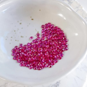May include: A white ceramic bowl filled with a cluster of small, faceted, hot pink gemstones. The bowl has a slightly irregular shape and sits on a marble-patterned surface. The gemstones are clustered in the center.