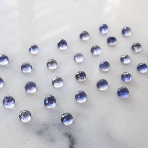 May include: A collection of small, faceted, purple gemstones arranged in a heart shape on a white surface.