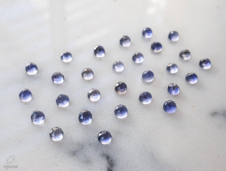 May include: A collection of small, faceted, purple gemstones arranged in a heart shape on a white surface.