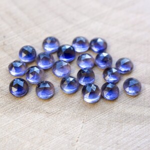 May include: A group of 17 faceted, round, purple gemstones. The gemstones are arranged in a loose cluster on a light brown wooden surface.