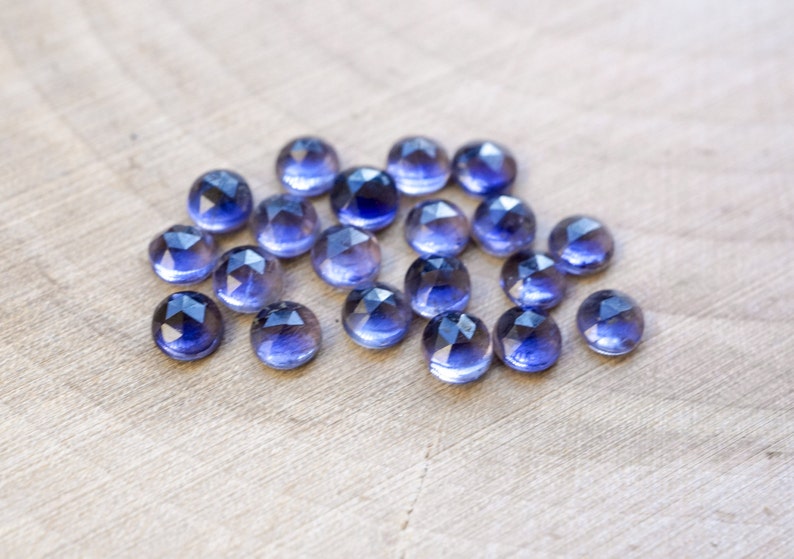 May include: A group of 17 faceted, round, purple gemstones. The gemstones are arranged in a loose cluster on a light brown wooden surface.