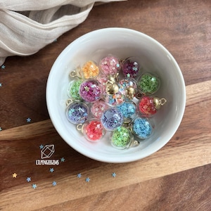 Op de afbeelding: Een verzameling kleine, doorzichtige glazen bolbedels gevuld met piepkleine, veelkleurige stervormige lovertjes. Elke bol heeft een goudkleurige metalen dop met een oogje om op te hangen. De bedels zitten in een witte kom, met verspreide sterlovertjes en de tekst LILPENGEEGEMS in het wit gedrukt.