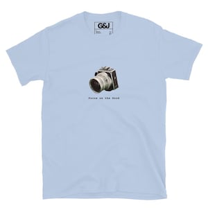 May include: A light blue short-sleeve t-shirt featuring a graphic of a vintage camera in the center of the chest. Below the camera, the text reads "Focus on the Good" in a typewriter-style font. The inner neck label displays the text "G&J".